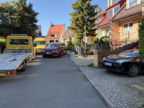 Residential street with parked cars, yellow vehicles on left, brick buildings with red-tiled roofs, trees and clear blue sky