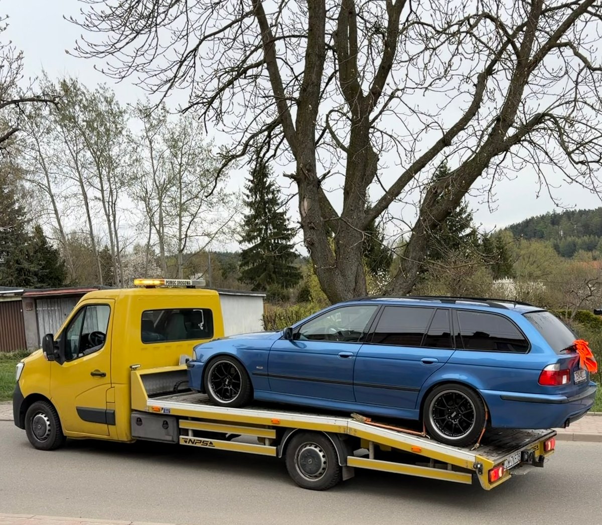 Yellow tow truck carrying a blue station wagon on its flatbed, with bare trees and rural buildings in the background