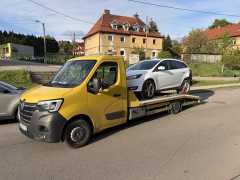 Yellow tow truck with flatbed transporting white SUV on residential street with European-style buildings in background