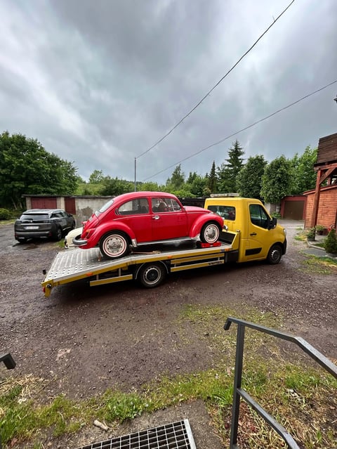 Red vintage Volkswagen Beetle being transported on a yellow tow truck in a rural storage yard